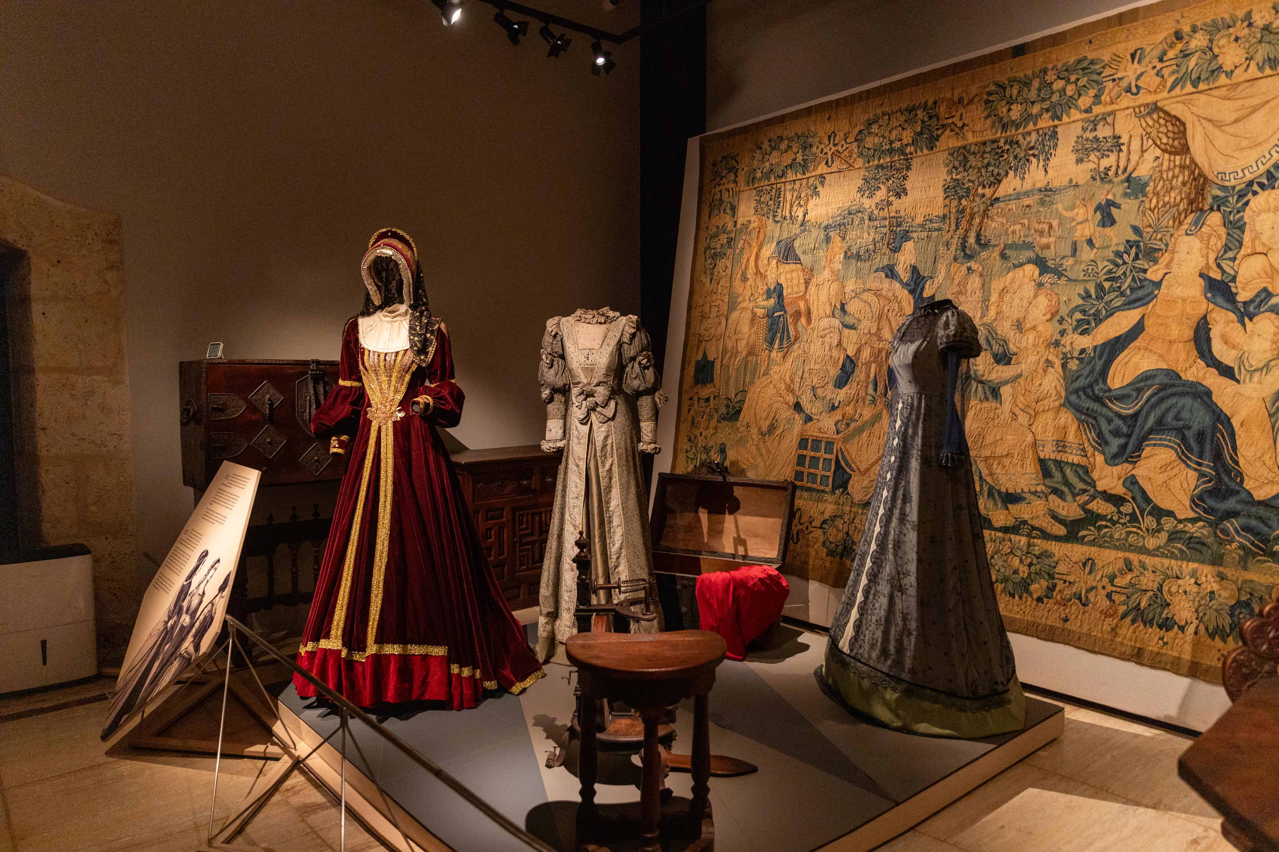 Sala de exhibición que muestra indumentaria histórica y trajes de época renacentista frente a un gran tapiz tejido con motivos florales y alegóricos.